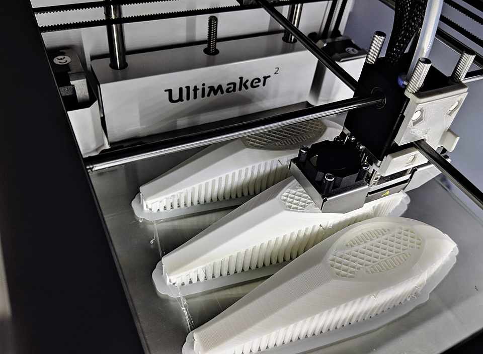 Ultimaker 3D printer mid-print, nozzle laying down a lattice-infill part with the drawer of finished parts below.