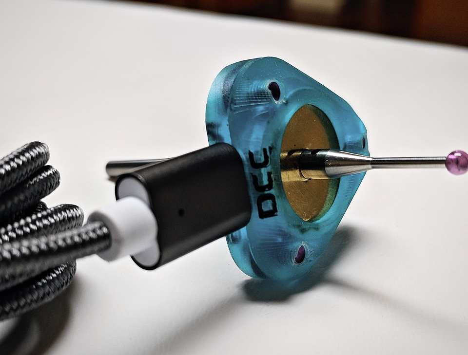 Macro shot of a teal 3D-printed micro-fixture with a steel pin probe through its center, held for scale on a workbench.
