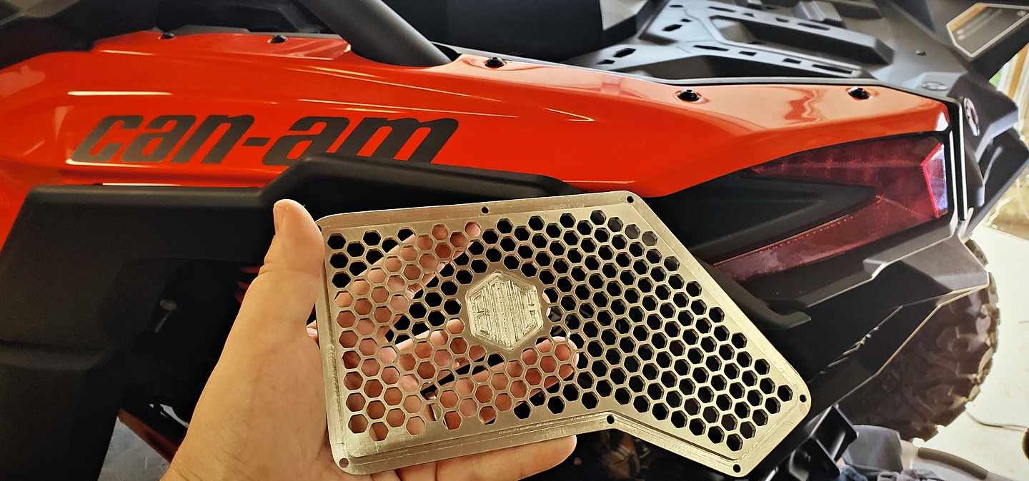 Holding a CNC-cut hexagonal-pattern intake grille up against the orange Can-Am Maverick X3's engine bay.