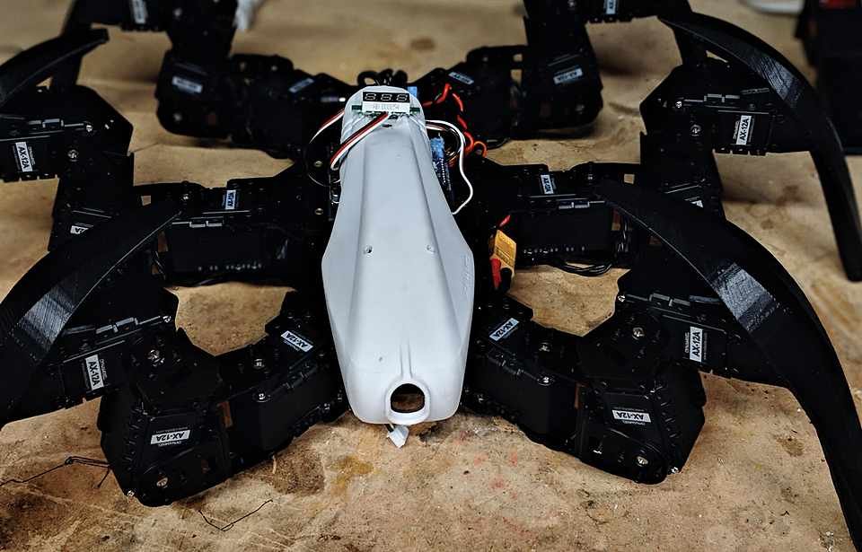 AX-12A servo hexapod robot on the workbench, six 3D-printed legs and a white sensor pod, wiring still exposed mid-build.